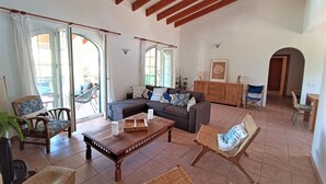A spacious living area with a sofa, chairs, a wooden coffee table, and a cabinet. Large windows provide natural light and a view of the outdoors. - Villa Persephone (Binibequer)