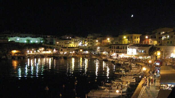 A nighttime harbor scene with boats docked along the waterfront, buildings with illuminated windows, and a walkway for pedestrians. - Villa Persephone (Binibequer)