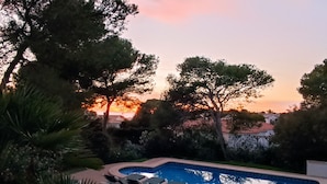 A swimming pool with lounge chairs, surrounded by trees and a sunset view. - Villa Persephone (Binibequer)