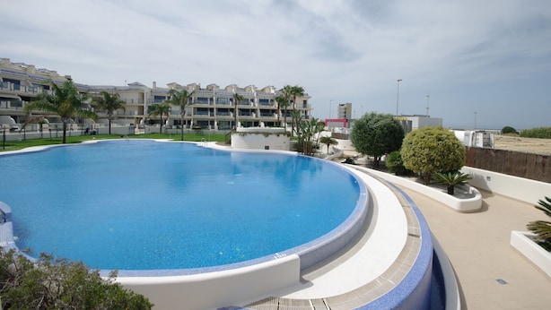 A large, circular swimming pool with a clear blue water surface, surrounded by a low white wall and a paved deck. In the background, there are multi-story buildings with balconies and palm trees. - 108 - Paraje de Los Lances (Tarifa)