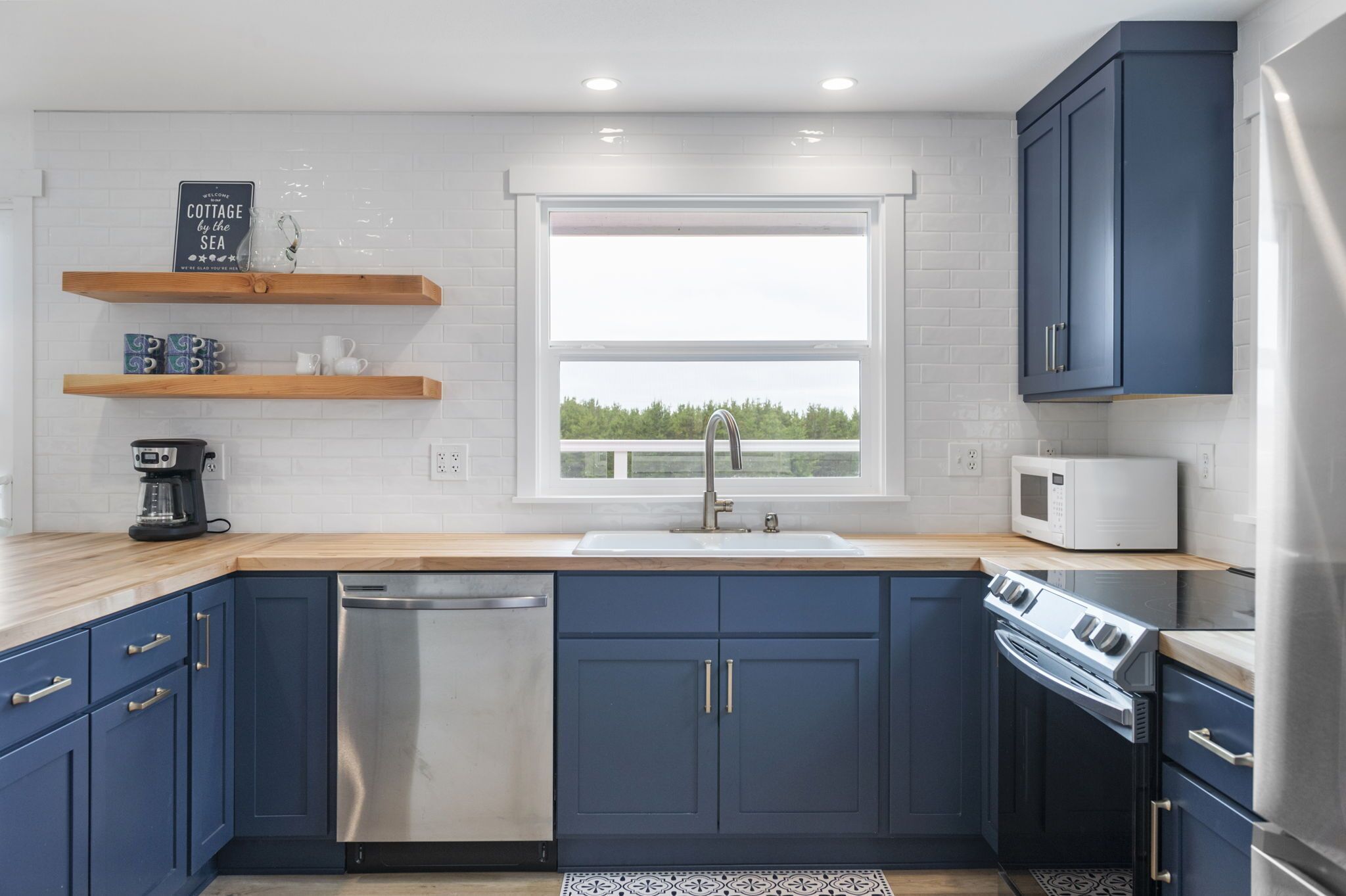 A modern kitchen with blue cabinets, a stainless steel dishwasher, a built-in oven, and a window with a view of trees.