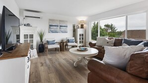 A modern living room with a brown leather sofa, a white coffee table, and a TV on a wooden cabinet. There is a fireplace with a mantel, a dining table, and a large window offering a view of trees. - HOT TUB! Sunset Views. Walk to Beach. Pet Friendly. Sleeps 10! (Westport)