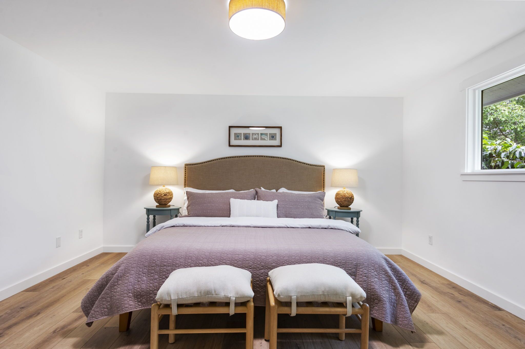 Une chambre avec un grand lit, deux tables de chevet avec des lampes, un tableau encadré au mur et une fenêtre donnant sur un paysage verdoyant.
