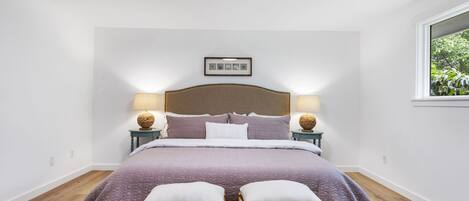 Une chambre avec un grand lit, deux tables de chevet avec des lampes, un tableau encadré au mur et une fenêtre donnant sur un paysage verdoyant.