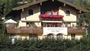 Un edificio de dos plantas con un balcón adornado con flores, un cartel que dice "APARTMENT Haus Jäger" y un paisaje de montañas al fondo.