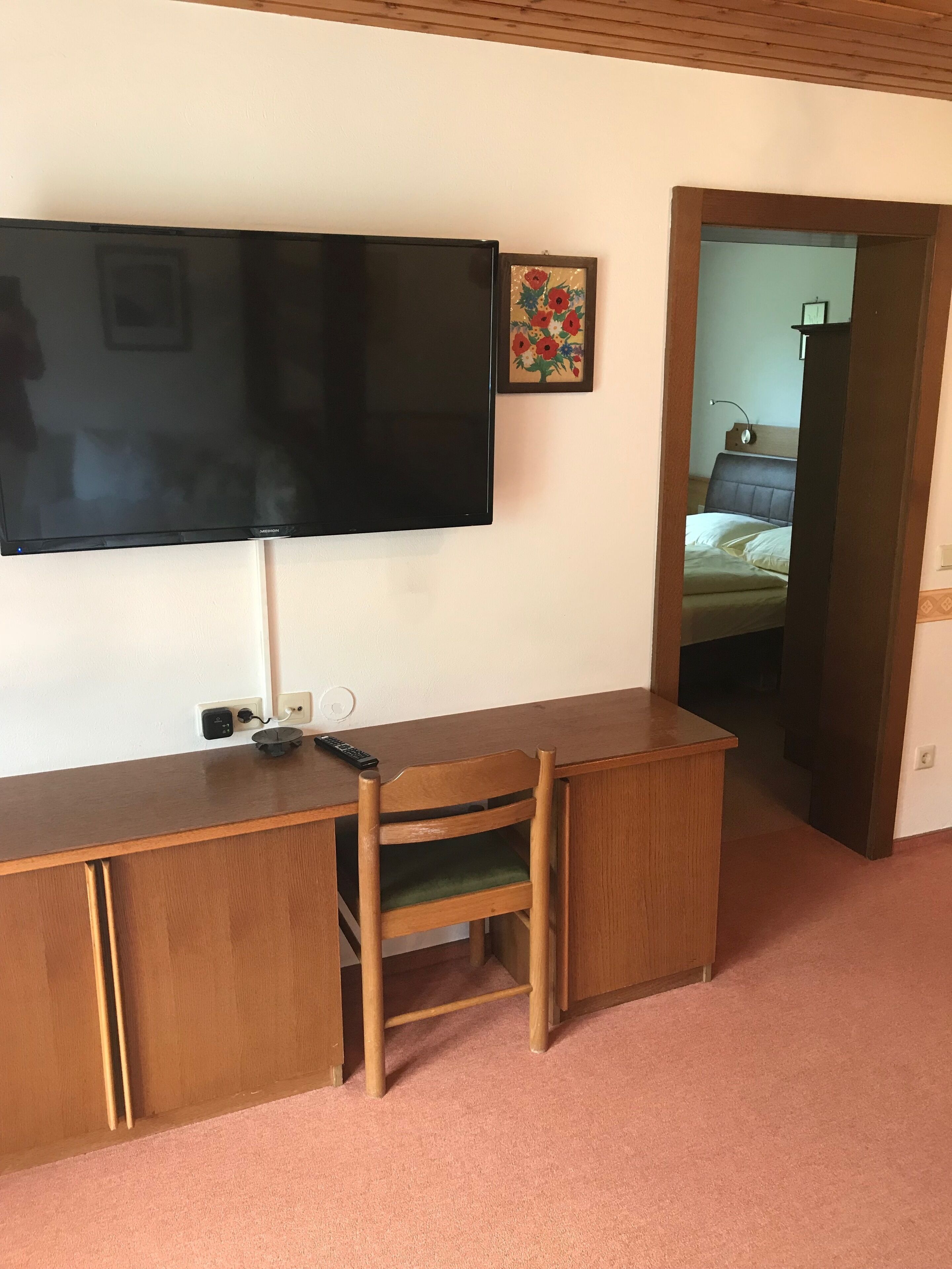 Une pièce avec un bureau en bois, une chaise, un téléviseur fixé au mur et un lit visible à travers une porte ouverte.