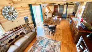 A cozy cabin interior with a wooden ceiling, a sofa with patterned cushions, a dining area with a table and chairs, and a kitchen with a stove and sink.