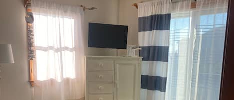 A bedroom with a bed, a ceiling fan, a TV, a dresser, and a window with curtains.
