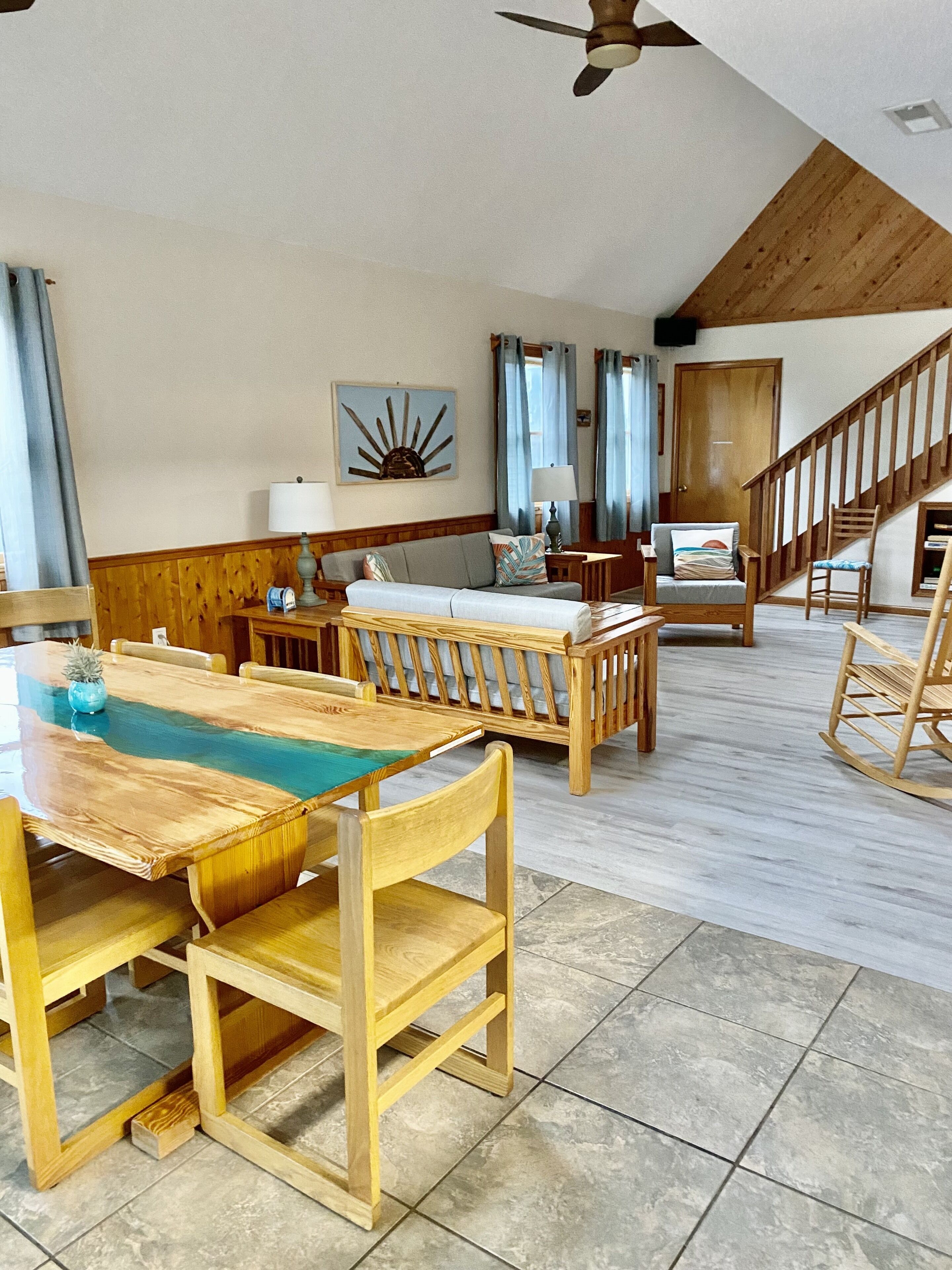 En rummelig opholdsstue med et træspisebord, en sofa og en gyngestol. En trappe fører op til en etage over.