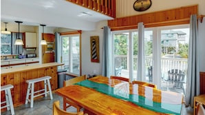 A wooden dining table with chairs, a kitchen area with a bar, and large windows with curtains. - Avon NC OBX Dog-Friendly Home  - Short walk to the Beach or Sound - PARK PLACE (Avon)