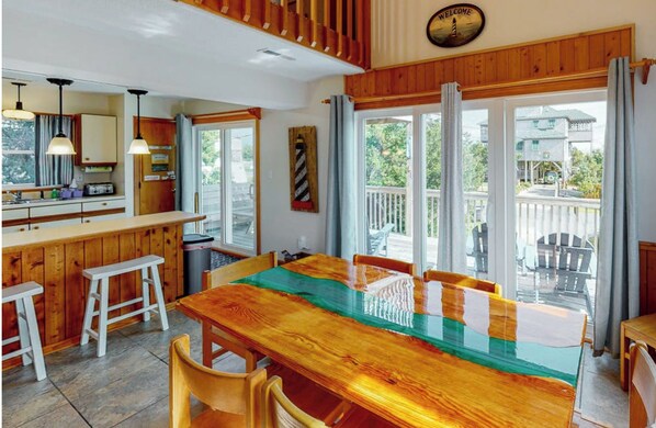 A wooden dining table with chairs, a kitchen area with a bar, and large windows with curtains.