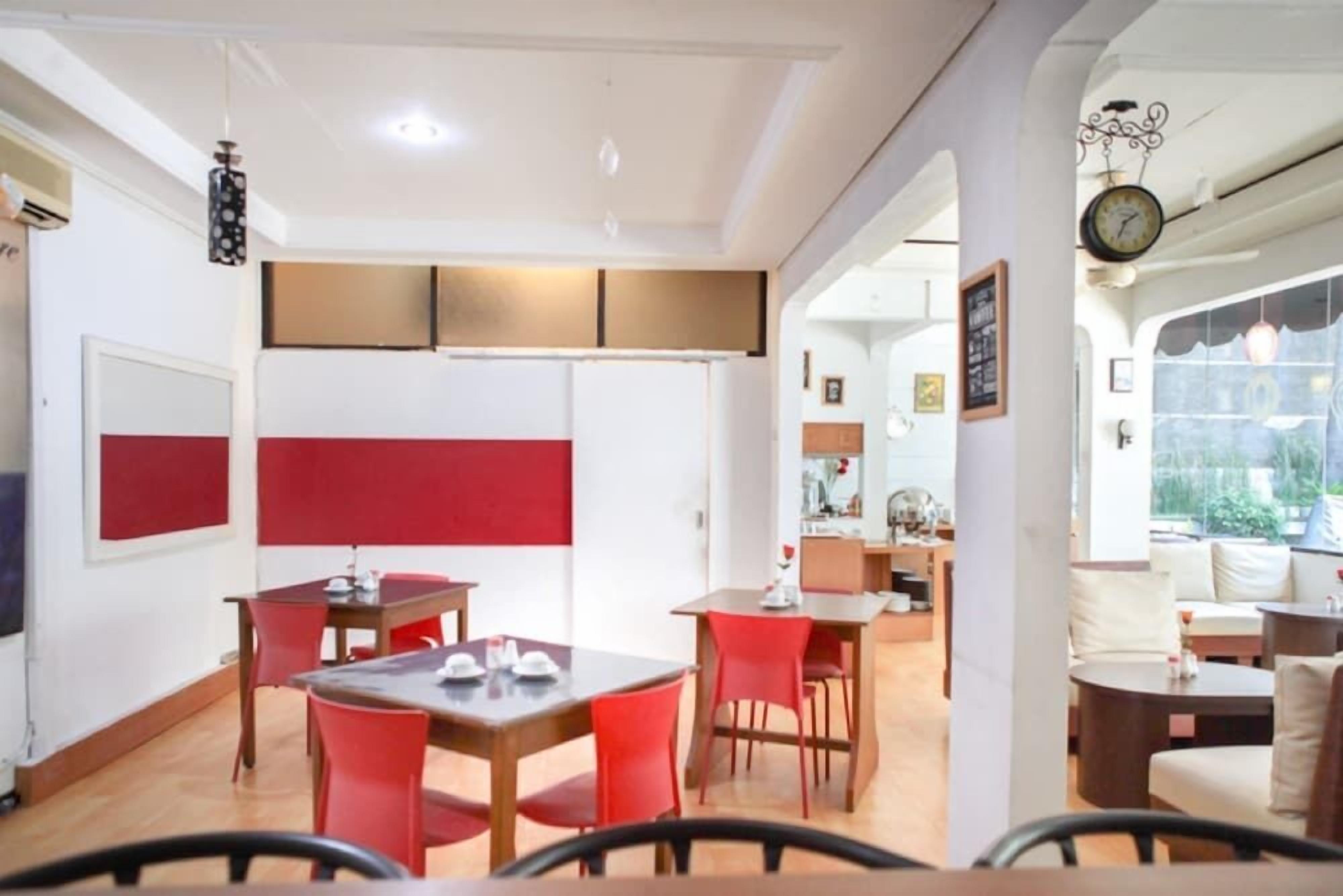 A dining area with red chairs and wooden tables, a kitchen in the background, and a clock on the wall.