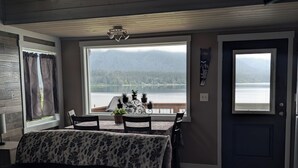 A room with a bed, a dining table with chairs, a view of a lake, and a door.