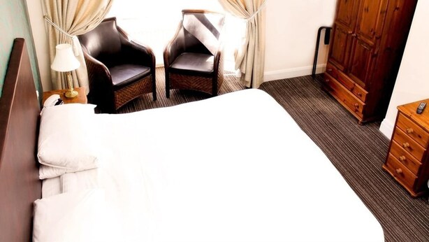 Una habitación de hotel con una cama grande, dos sillones, un armario de madera y una cómoda pequeña de madera.