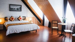 Een slaapkamer met een schuine balkenlaag, een bed, twee stoelen, een tafeltje en een raam met gordijnen.