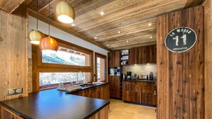 A modern kitchen with wooden cabinetry, a central island, pendant lighting, and a view of the outdoors through a large window. - Chalet ski in / out avec vue, emplacement fabuleux (Meribel Les Allues)