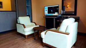A hotel room with two white leather armchairs, a small wooden table, a flat-screen TV, a mirror, and a desk with a lamp.