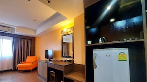 A hotel room with a wooden floor, an orange chair, a desk with a television, a mirror, and a refrigerator.