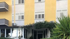 A hotel entrance with a stone pathway leading to a covered area, a bar, and a well-maintained garden with a palm tree. - Hotel Sabatino (Follonica)