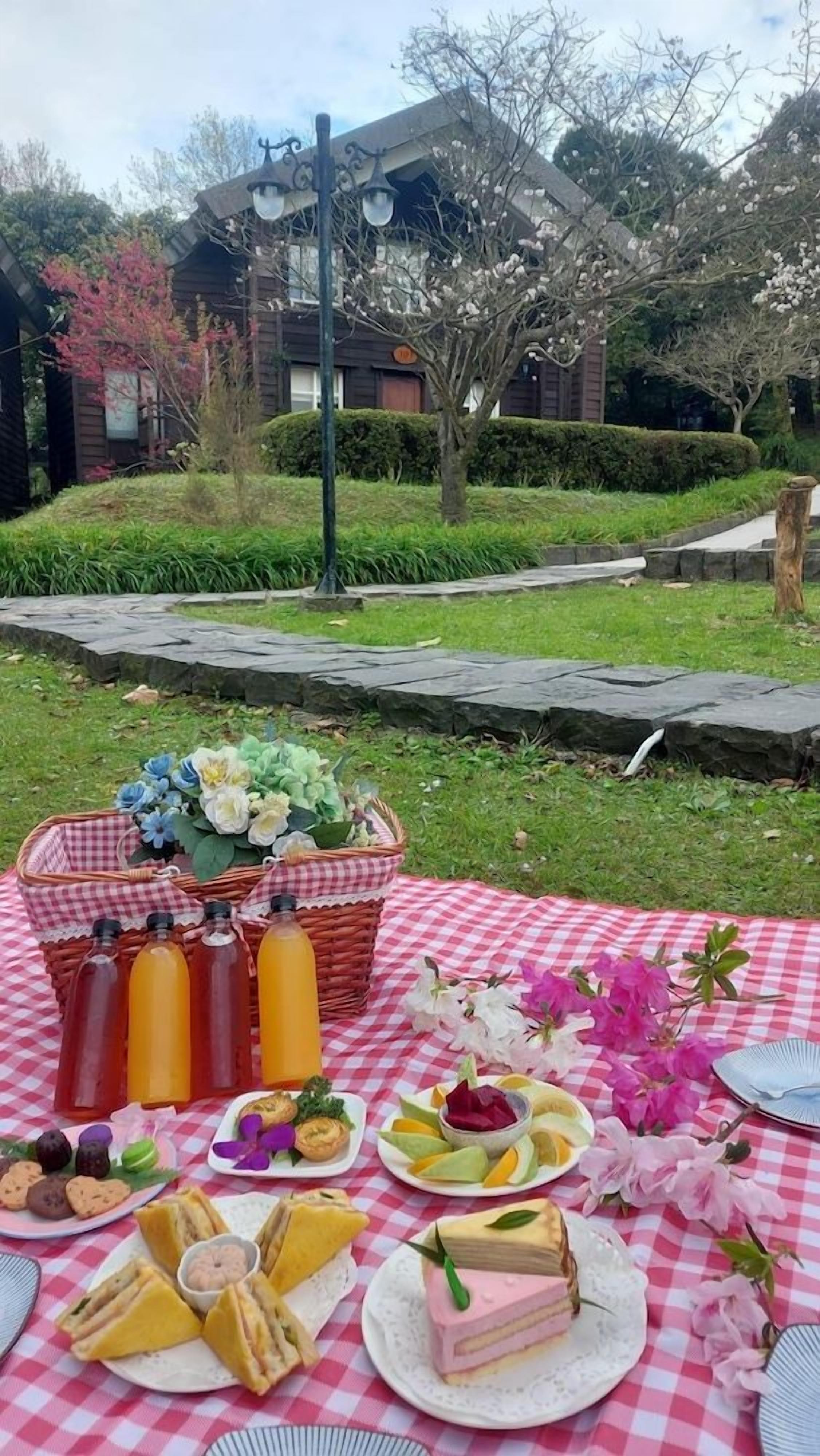 A picnic setup with a checkered tablecloth, a basket of flowers, and various food items.
