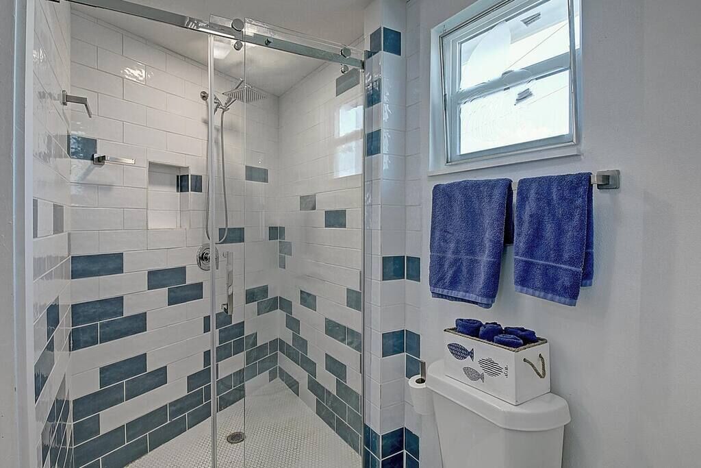 Ein Badezimmer mit einer gläsernen Duschkabine, weißen und türkisfarbenen Fliesen, einer Toilette und zwei blauen Handtüchern, die auf einem Haken/Gestell hängen.