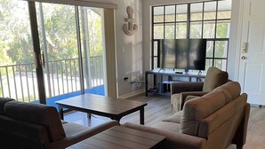 A spacious living room with a large TV, a ceiling fan, and a balcony with a view of trees. - Manatee Manor. Waterfront, dock, stilted home. Renovated June '23.  (Homosassa)