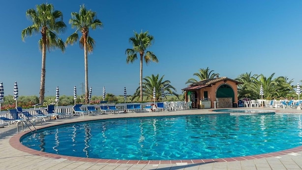 A swimming pool with lounge chairs and umbrellas, surrounded by palm trees and a building with a red roof. - Agrihotel Elisabetta (Cecina)
