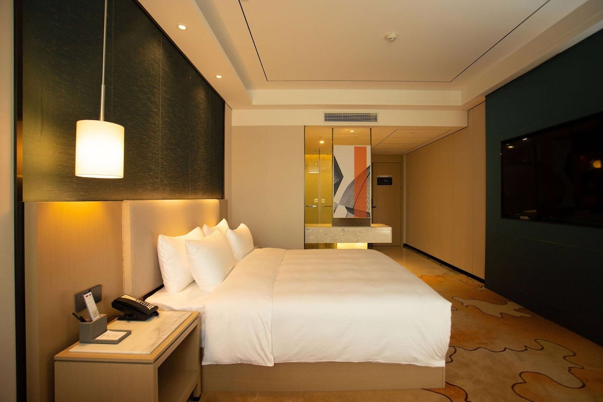 Habitación de hotel con una cama grande, una mesita de noche con lámpara y vistas al pasillo.