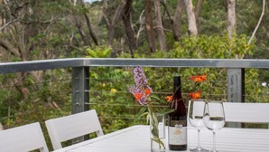 Una mesa con una botella de vino, copas y un jarrón con flores en un balcón con vistas a árboles.