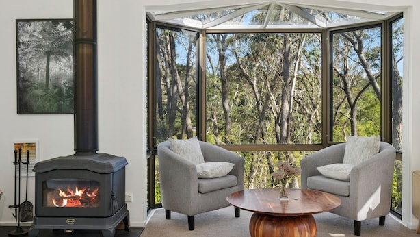 Un salón acogedor con una estufa de leña, dos sillones grises, una mesa de centro redonda de madera y un ventanal con vistas a un bosque.
