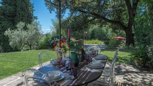 Un área de comedor al aire libre con una mesa preparada para seis personas, rodeada de sillas, en un entorno de jardín con árboles y una sombrilla roja al fondo.