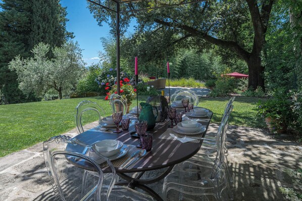 An outdoor dining area with a table set for six, surrounded by chairs, in a garden setting with trees and a red umbrella in the background.