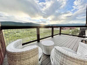 Une terrasse en bois avec des chaises en osier et une vue sur un champ verdoyant et des montagnes.