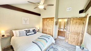 A bedroom with a large bed, a ceiling fan, wooden doors, a bathroom with a mirror, and a nightstand with a lamp.