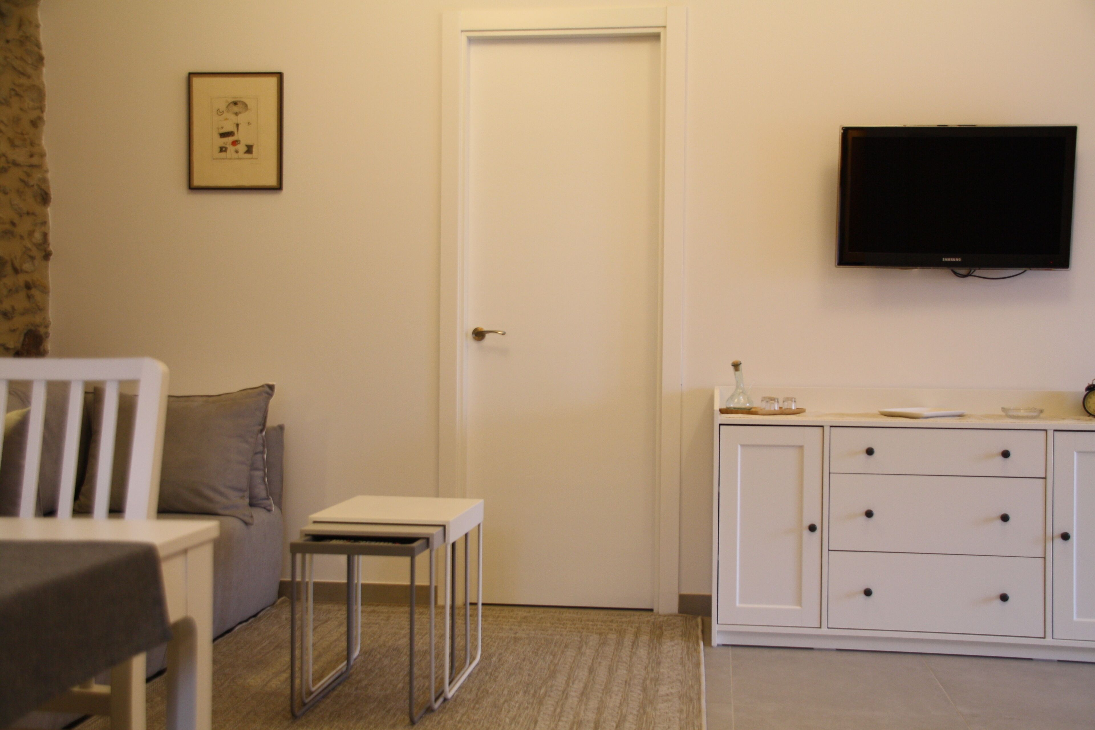 Une pièce avec une porte blanche, un téléviseur à écran plat fixé au mur, une petite table blanche et un meuble blanc avec des tiroirs.
