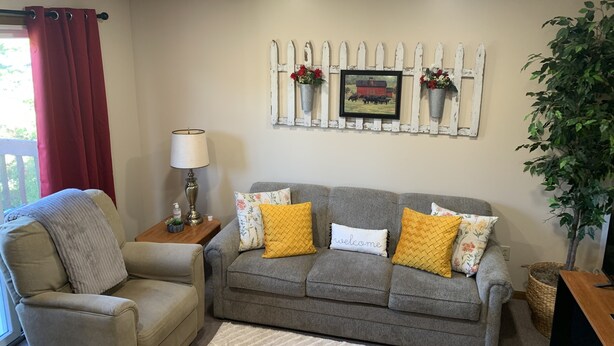 Un salon avec un canapé gris, un fauteuil beige et une petite table sur laquelle se trouve une lampe. Un mur décoratif est agrémenté d’une clôture blanche en bois et d’un tableau encadré.