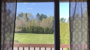 Une vue par la fenêtre, à travers des rideaux à motifs, donnant sur une pelouse et des arbres.