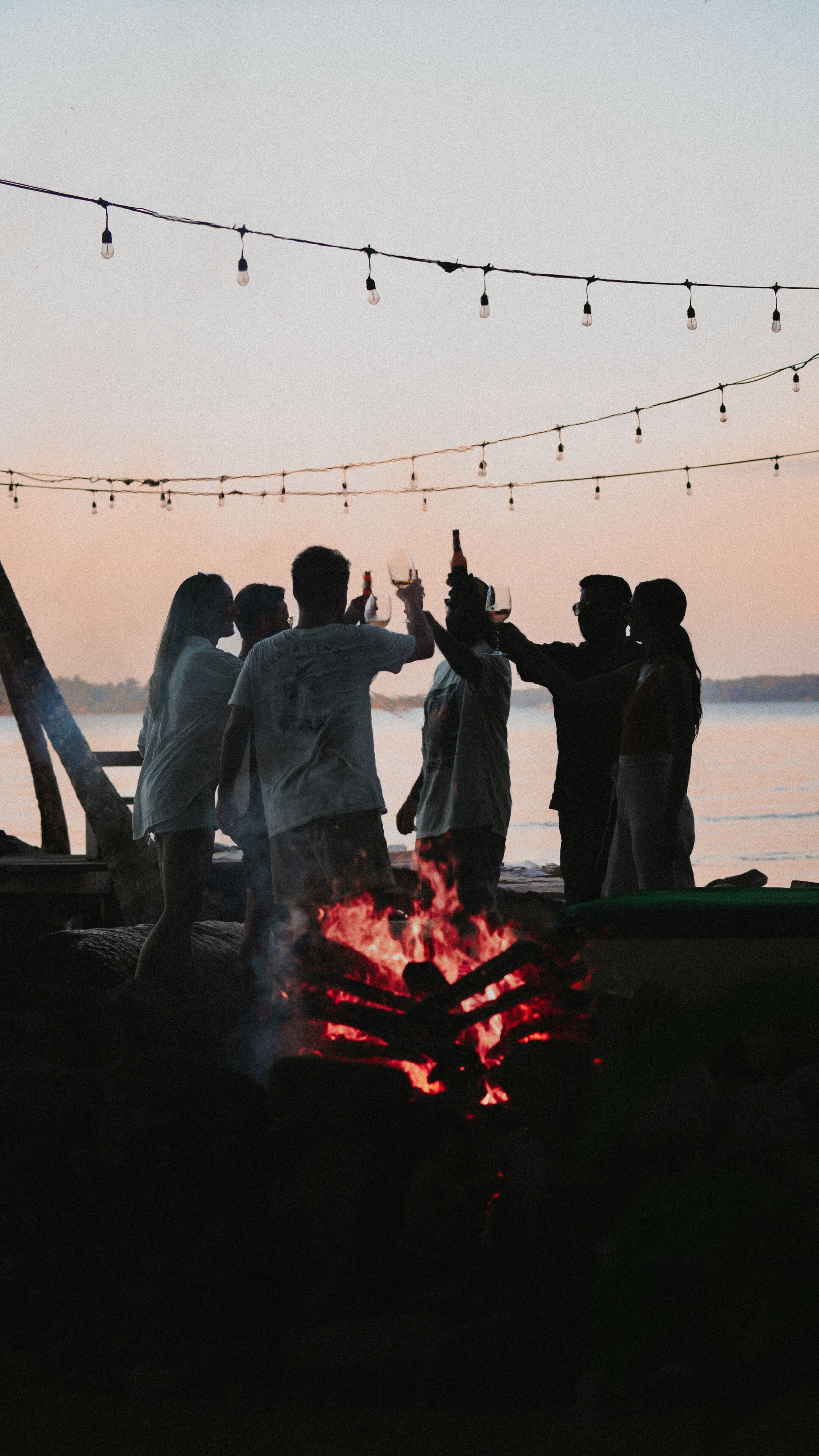 Un grupo de personas alrededor de una fogata al anochecer, con luces colgantes sobre ellas.