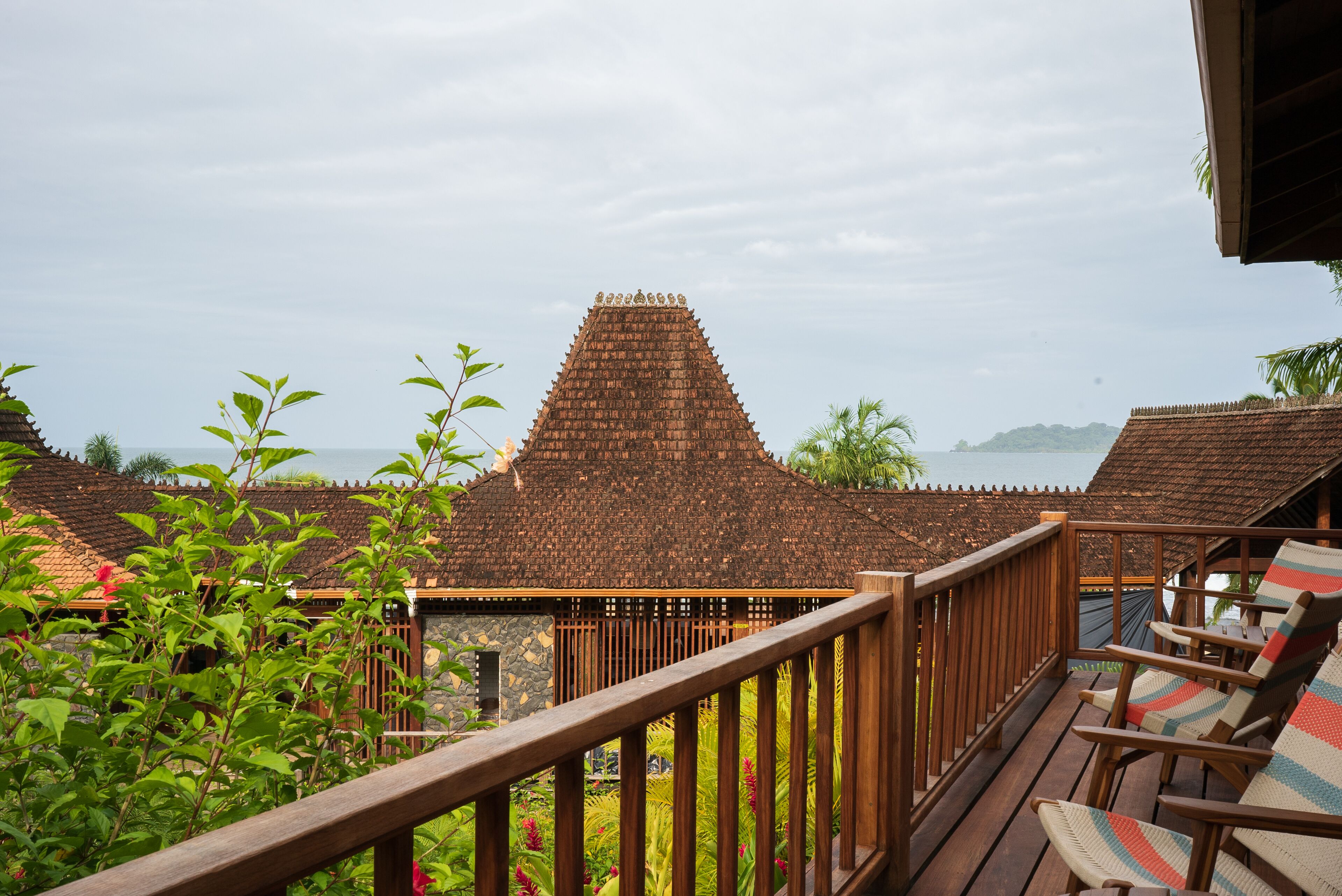 A wooden deck with a railing, overlooking a traditional building with a tiled roof and a view of the sea.
