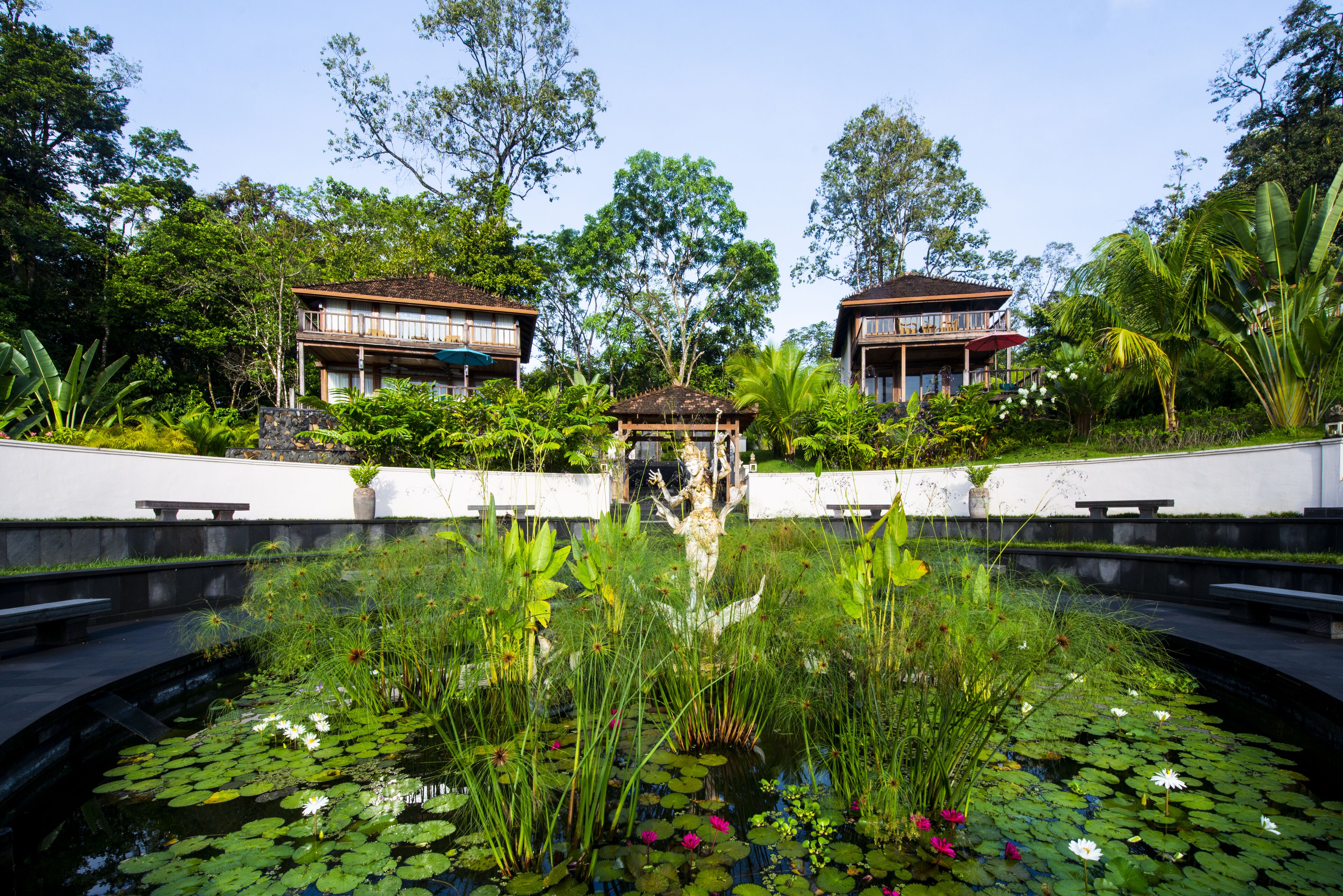 Un jardín tranquilo con un estanque, dos casas de madera y abundante vegetación.