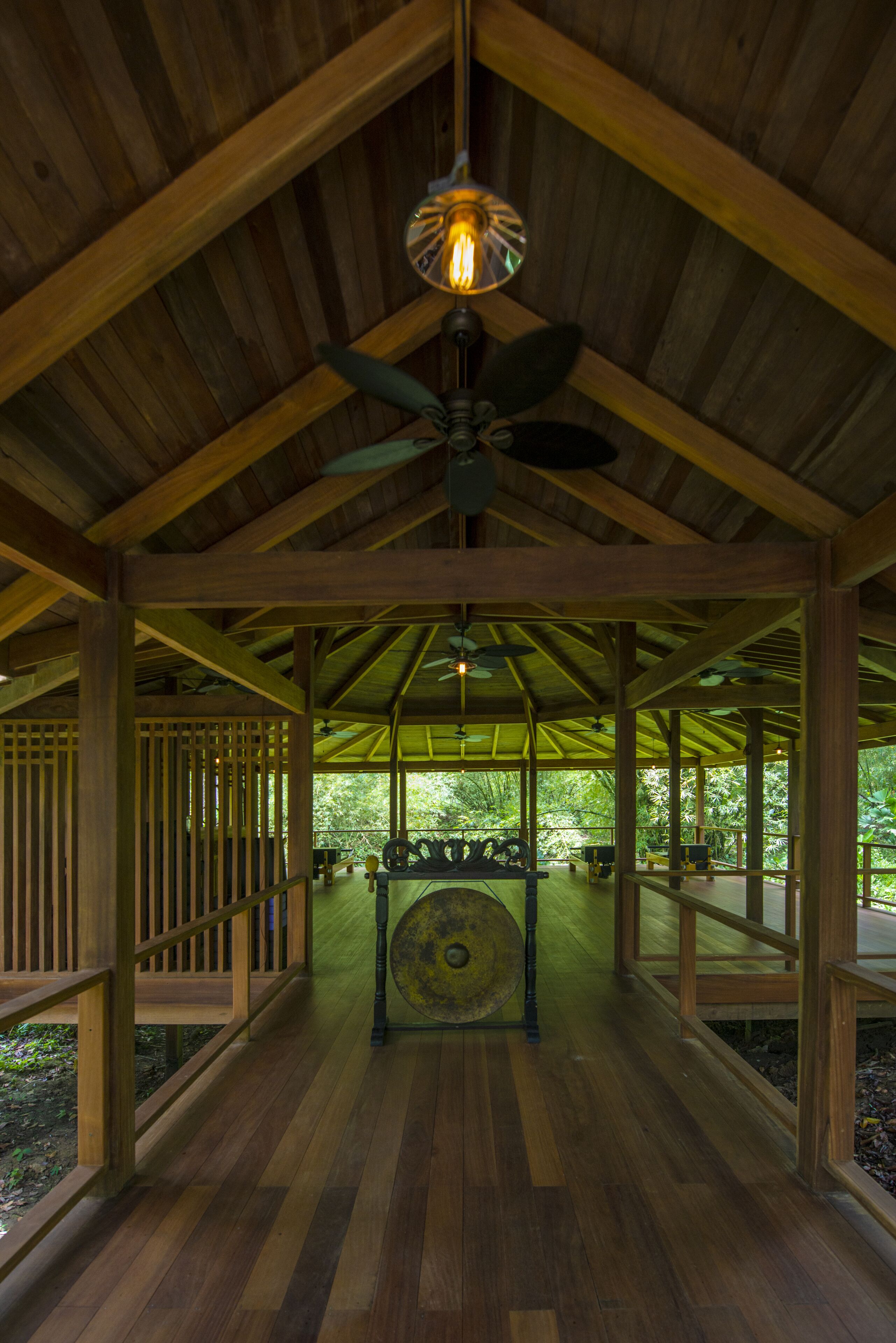 A covered wooden pavilion with a central seating area, ceiling fans, and a view of the surrounding greenery.