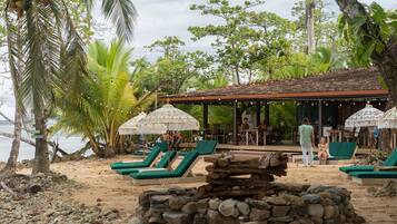 Un resort frente al mar con sillas de descanso, una estructura de techo de paja y un pozo de fuego de piedra.