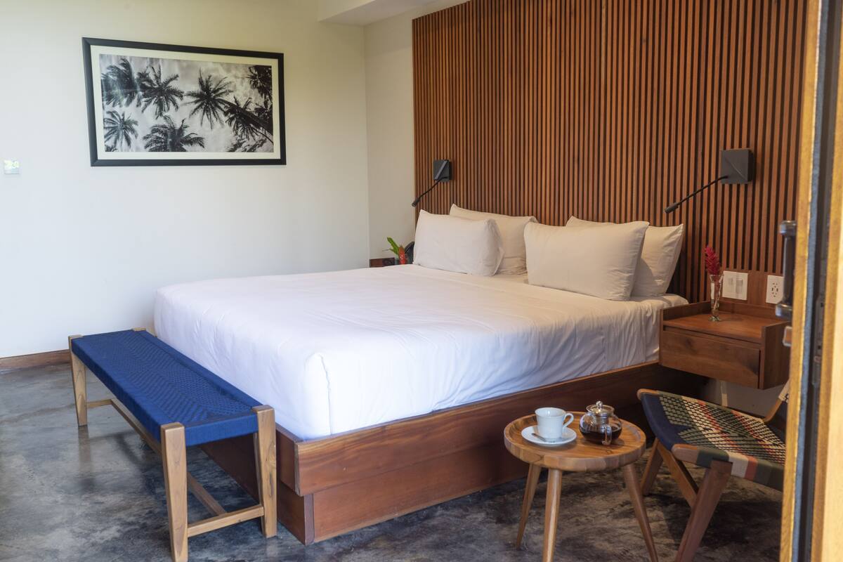 A neatly arranged bedroom with a large bed, a bench, a small table with a cup and a teapot, and a wall-mounted lamp.