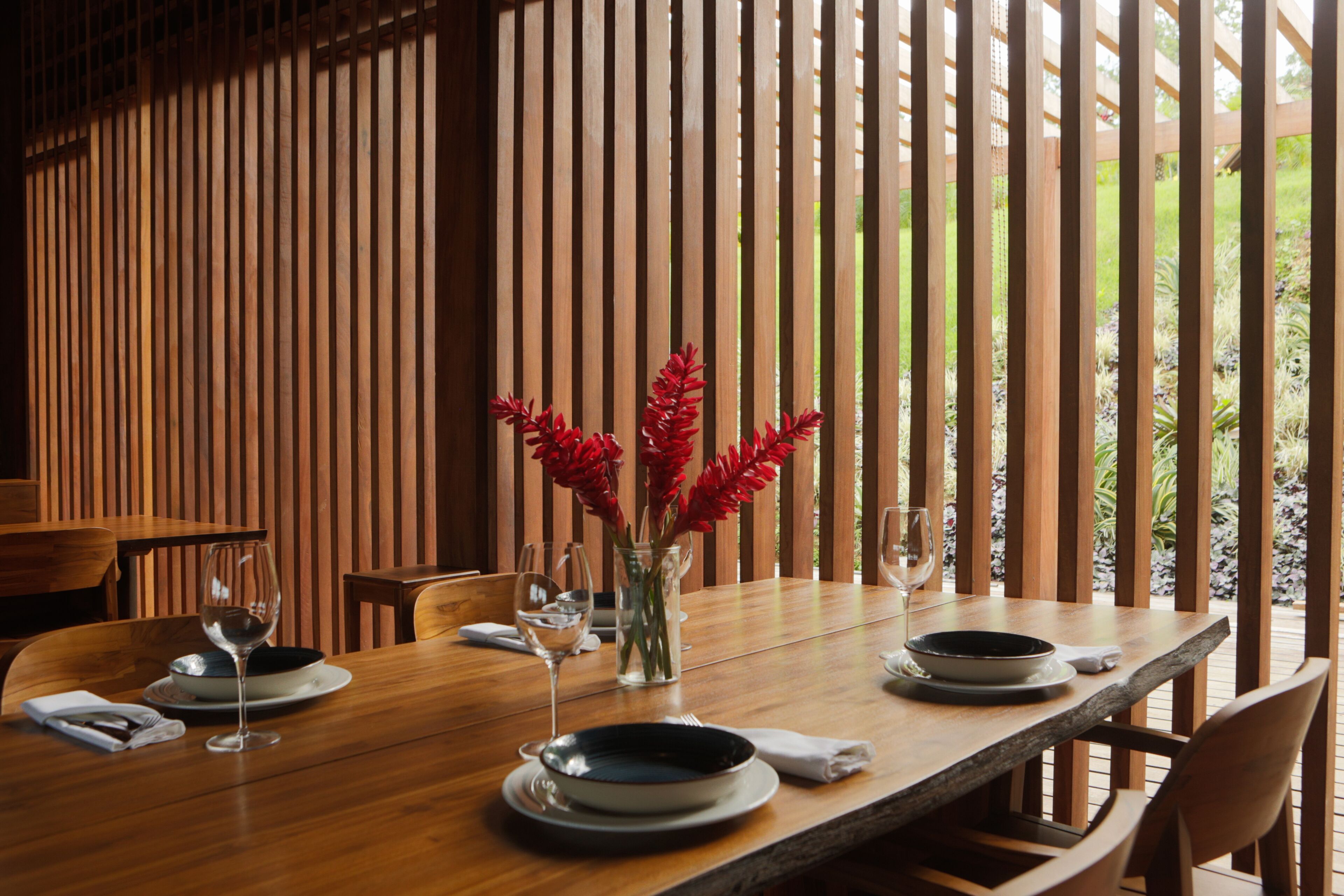 Una mesa de madera con platos, vasos y un jarrón de flores rojas.
