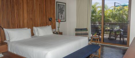 A bedroom with a large bed, wooden headboard, a bench, a small table with a vase, and a view of a patio with outdoor furniture.