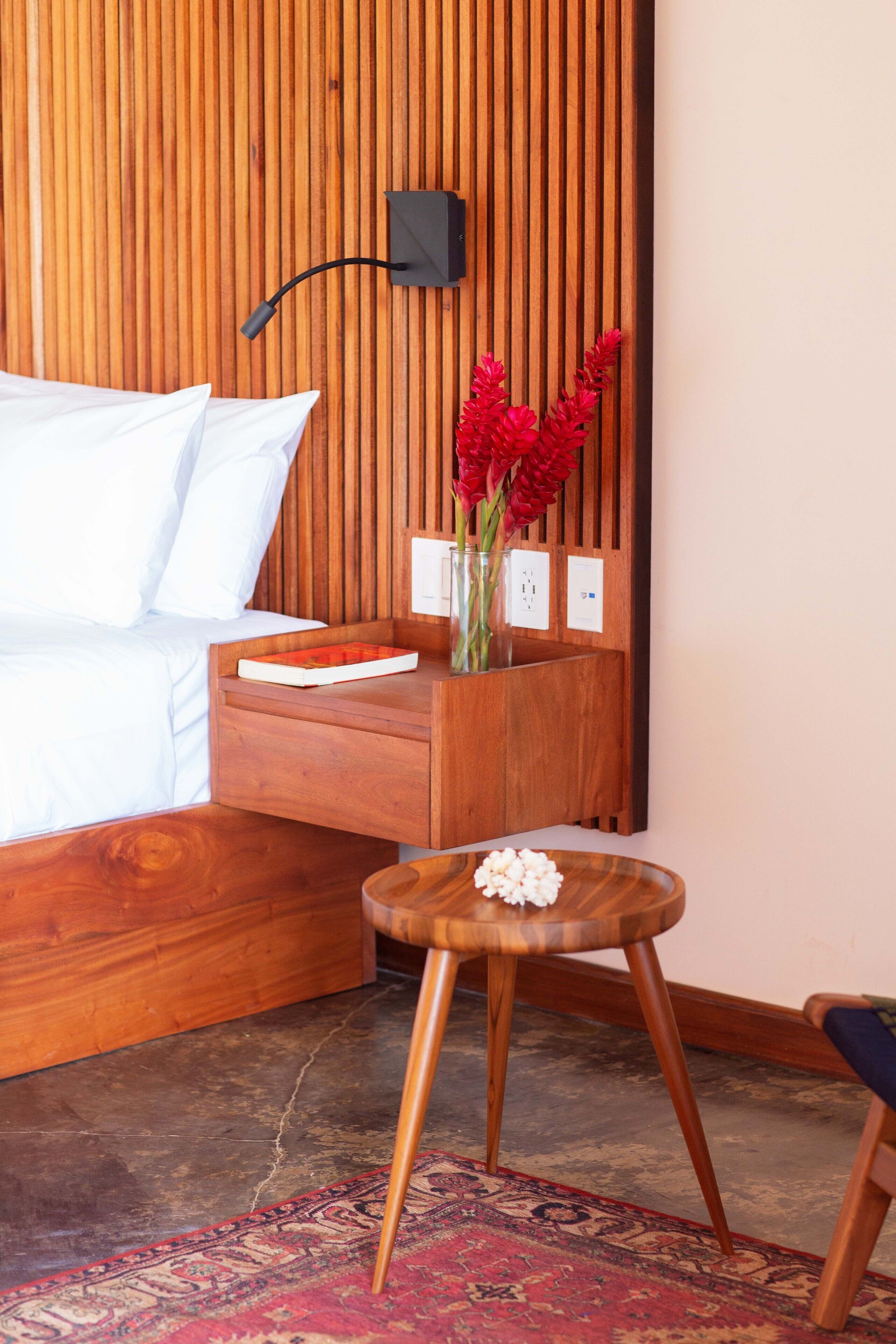 Habitación de hotel moderna con cabecera de madera, mesita de noche con un jarrón de flores rojas, un taburete pequeño de madera y una alfombra roja con estampado.