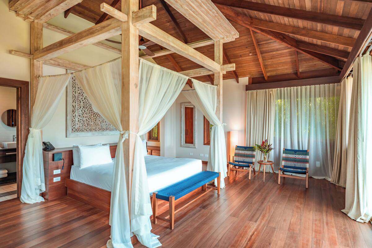 A spacious room with a wooden ceiling, a large bed with a canopy, a bench, two chairs, and a small table with a vase of flowers.