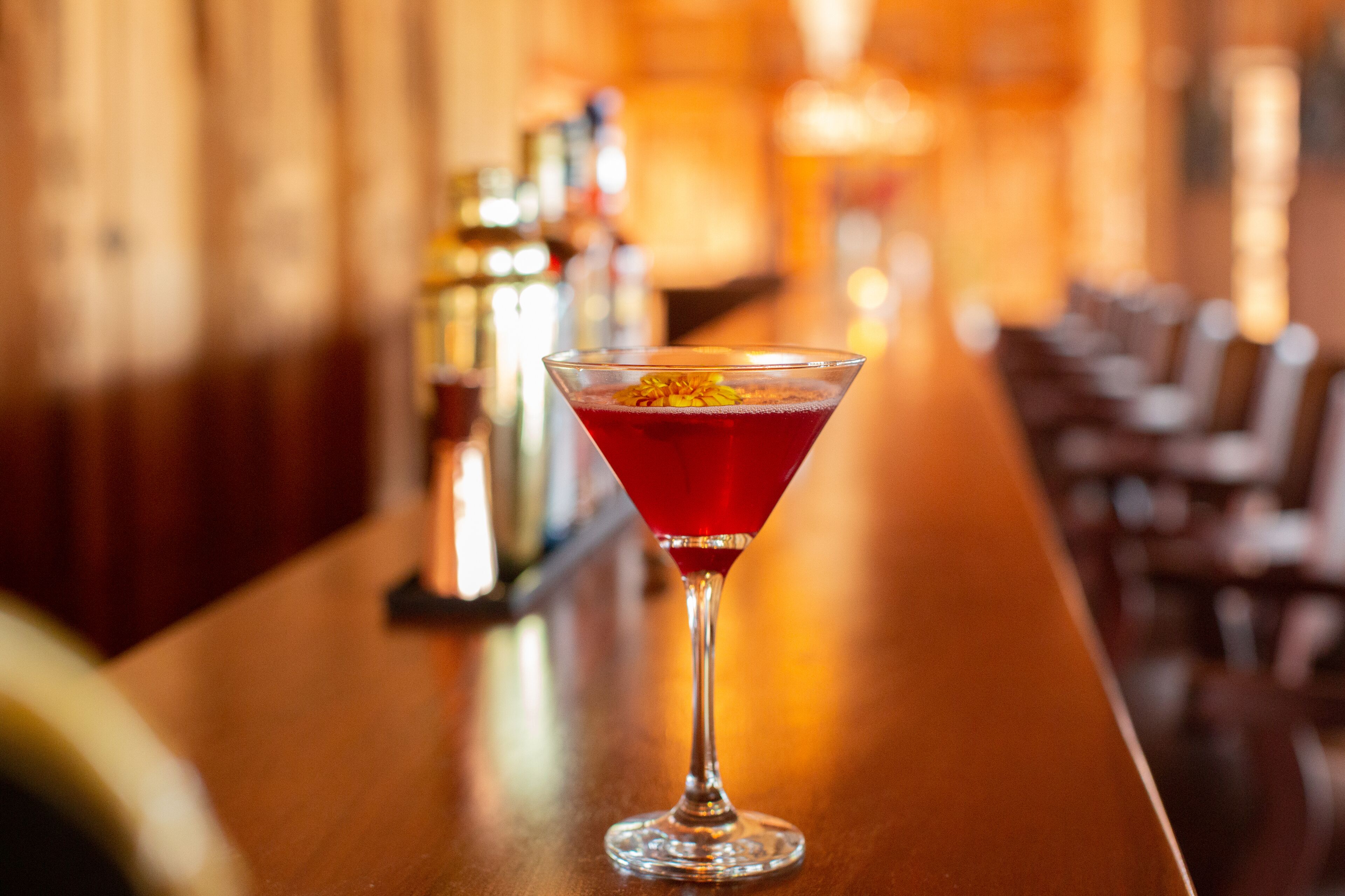 A bar setting with a martini glass containing a red drink, a bar counter, and a blurred background with bar stools.