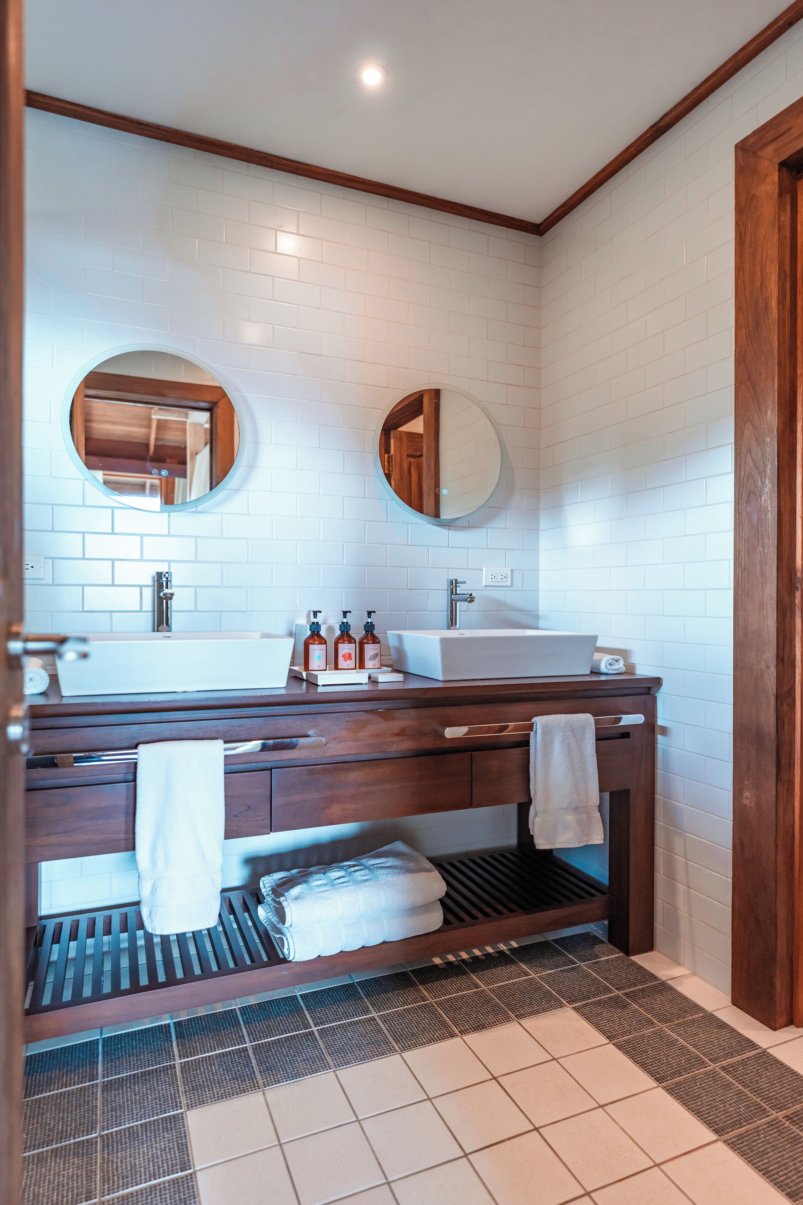 A bathroom with a white tiled wall, wooden vanity, two round mirrors, a white sink, and a towel rack.