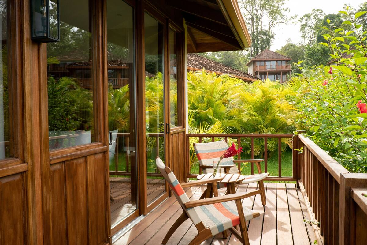 A wooden deck with two lounge chairs, a glass door leading to a lush garden, and a wooden house in the background.